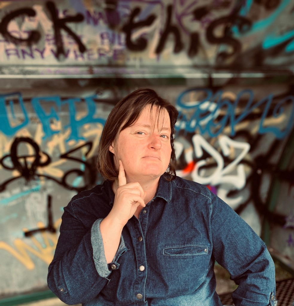 Ginny Evans-Pollard posing in front of colourful graffiti at a Bristol shelter, with one hand raised beside the words “Fuck This.”